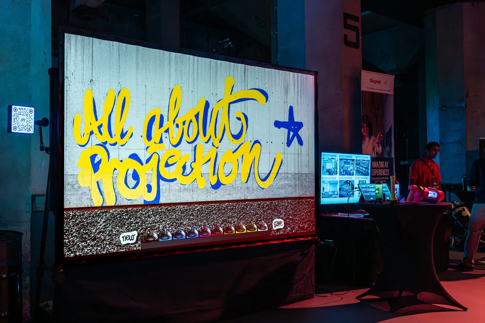 All About Projection in licht gespoten over een geprojecteerde betonnen muur, met een rek spuitbussen eronder